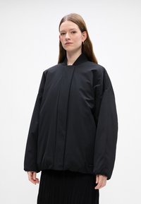 Zwarte gewatteerde jas met een hoge kraag, oversized pasvorm en elastische manchetten. Zachte textuur met een voorste plooi-detail en een logo-label aan de zijkant.