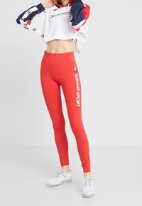 Donna che indossa leggings rossi a vita alta con il logo "TOMMY SPORT" e una felpa corta bianca, rossa e blu navy, scarpe da ginnastica bianche, in posa all'interno.