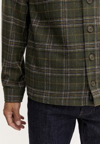 Roheline flanellshirt plaidi mustriga, millel on pruun, sinine ja valge kombinatsioon. Omab nööpide kinnitusi ja pehmet tekstuuri. Kanda tume teksadega.