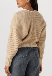 Vrouw in een beige chunky gebreide trui met ballonmouwen en high-waisted vervaagde zwarte jeans, gezien vanuit de rug, tegen een effen achtergrond.