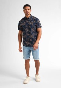 Camicia a maniche corte blu navy con un motivo floreale beige, abbinata a pantaloni corti di denim chiaro. Il modello indossa sneakers bianche.