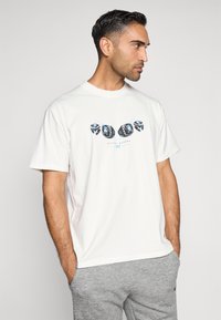 T-shirt de algodão branca com mangas curtas, apresentando um design gráfico de elementos pretos e azuis na parte da frente, combinada com calças de moletom cinzentas.