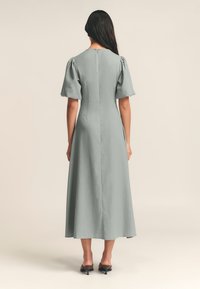 Abito lungo a forma A in tessuto verde tenue, con maniche a sbuffo, scollatura alta e chiusura con zip sul retro; indossato con sandali neri con tacco.