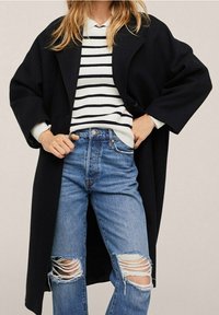 Manteau noir surdimensionné avec fermeture à boutons, porté sur un pull rayé blanc et marine, associé à un jean bleu déchiré aux genoux.