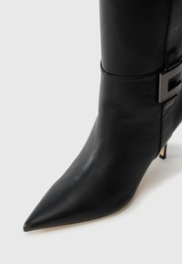 Black leather ankle boot with a pointed toe, sleek design, and a small heel. Features a side strap with a metallic buckle detail.