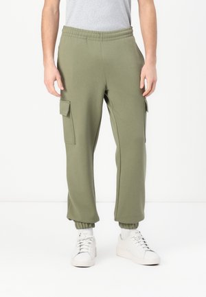Hombre vistiendo pantalones jogger cargo verde oliva con puños elásticos y zapatillas blancas, de pie contra un fondo blanco liso.