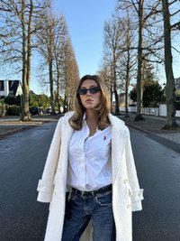 Weißer Wollmantel mit großen Knöpfen, weißes Hemd, dunkle Jeans und schwarze Sonnenbrille. Vor einer von Bäumen gesäumten Straße.