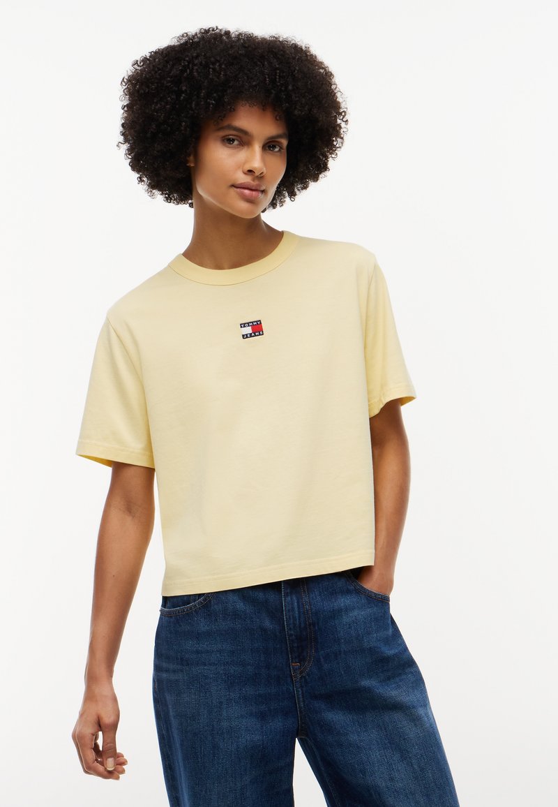 Jeune personne aux cheveux bouclés porte un t-shirt jaune pâle à manches courtes avec un petit logo et un jean bleu sur un fond blanc uni.