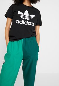 Černé tričko s bílým logem "adidas" a designem trojlístku, doplněné zelenými tepláky s kontrastními panely a logem.