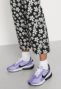 Piernas con pantalones negros con patrones de flores blancas, calcetines blancos y zapatillas Nike moradas con el logo de swoosh negro sobre un suelo gris.
