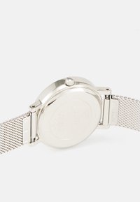 Silverfärgad klocka med rund boett, kedjeband och en krona. Slät metallisk yta på baksidan av boetten, minimalistisk design utan utsmyckningar.