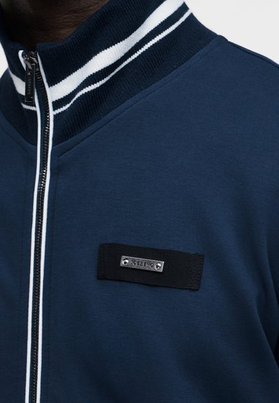Veste zippée bleu marine avec col côtelé comportant des rayures blanches et un patch noir avec une étiquette métallique "ICEBERG" sur la poitrine.