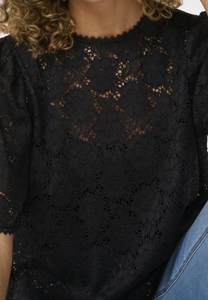 Vrouw draagt een zwarte kanten top met bloemenpatronen en geschulpte randen, gecombineerd met een blauwe spijkerbroek, tegen een witte achtergrond.