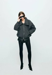 Czarna, oversize bomberka z dużym kołnierzem, zapinana z przodu na zamek błyskawiczny, w połączeniu z czarnymi szortami i rajstopami, uzupełniona ciemnymi okularami przeciwsłonecznymi.