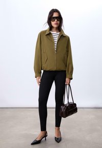 Olivgrüner Reißverschlussjacke mit zwei Seitentaschen, schwarzen, figurbetonten Hosen, schwarzen Slingback-Absätzen und einer kleinen braunen Handtasche. Gestreiftes Oberteil darunter.