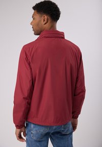 Veste rouge légère avec col montant et poignets élastiques. Texture lisse, coutures visibles et un design simple au dos.