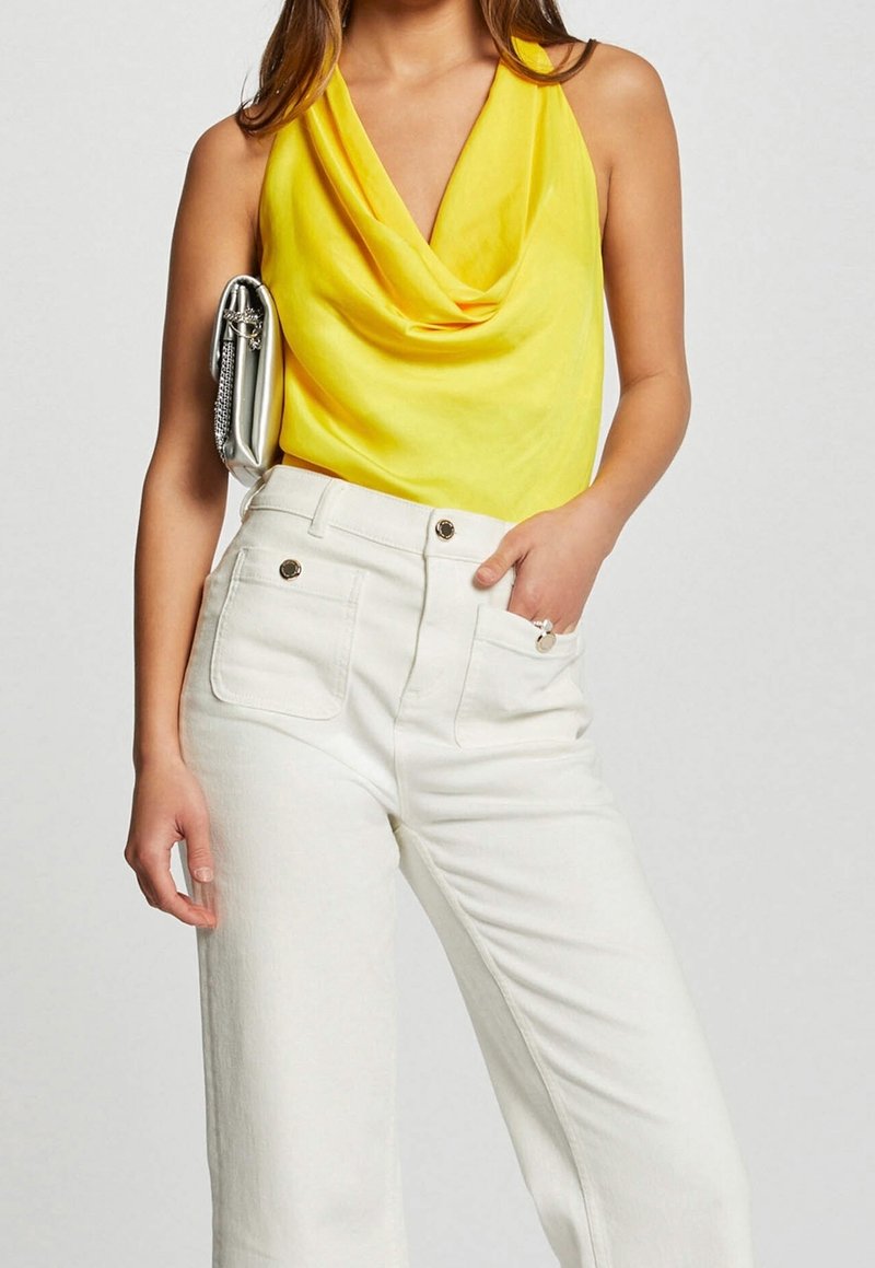 Haut jaune à col cheminée en tissu léger, associé à un pantalon large blanc taille haute avec des boutons métalliques et des poches.