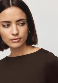 Brown top with a smooth texture, featuring a crew neckline and a slightly gathered shoulder detail. Model has shoulder-length dark hair.