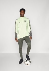Lys grønn zip-up treningsgenser med mørkegrå detaljer, parret med slim-fit mørkegrå bukser og svarte fotballsko, stående på en fotball.