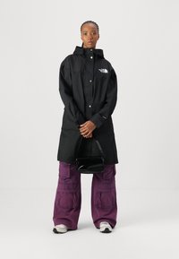 Trench noir avec une fermeture à pression à l'avant, logo sur la poitrine, associé à un pantalon cargo violet à coupe ample et des baskets blanches. Sac à main noir.