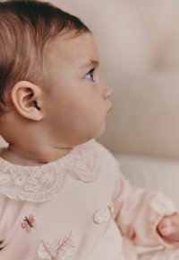 Prenda de color rosa claro con cuello de encaje; presenta bordados de mariposas y detalles florales. Perfil de un niño sobre un fondo suave y neutro.