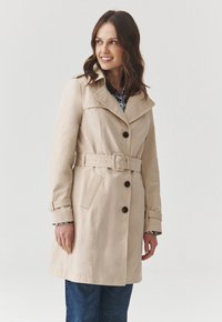Béžový trench coat s širokým límcem, zapínáním na knoflíky, pásem v pase a kapsami. Tkanina vypadá hladce s mírným leskem.