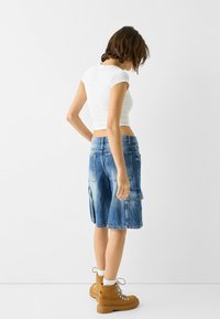 Shorts cargo en denim avec une finition délavée, des poches latérales et une coupe décontractée, associés à un crop top blanc ajusté et des bottes à lacets marron.