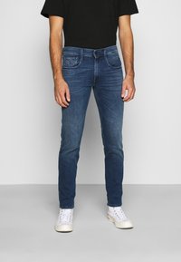 Jeans de mezclilla azul con un corte slim, diseño de cinco bolsillos y lavado desgastado, combinados con zapatillas blancas y una camiseta negra.