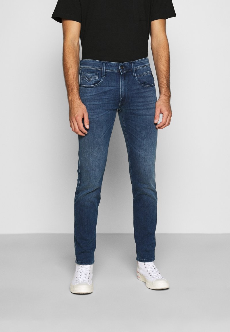 Jeans de mezclilla azul con un corte slim, diseño de cinco bolsillos y lavado desgastado, combinados con zapatillas blancas y una camiseta negra.