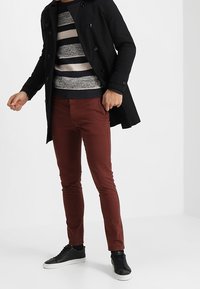 Mężczyzna w czarnym płaszczu, w paskowanym swetrze, rudych spodniach i czarnych sneakersach stoi na tle białej �ściany.