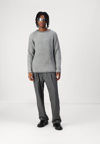 Pulover gri cu detalii în relief, cu mâneci lungi, asortat cu pantaloni gri și pantofi negri. Accesoriile includ ochelari de soare reflectivi. Fundal simplu.