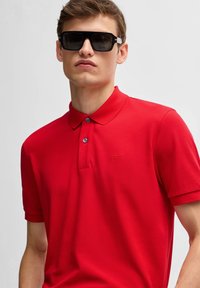 Röd polo skjorta i strukturerad tyg, med klassisk krage, två knappar och en liten broderad "BOSS"-logotyp på vänster bröst.