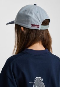 Ljusblå basebollkeps med marinblå skärm, med röd "hummel SPORTSWEAR" logotyp på baksidan. Bärs med en marinblå skjorta.