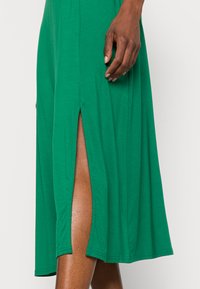 Vestido verde, até ao joelho, feito de um tecido macio, com fendas laterais e um corte descontraído. Detalhes de costura visíveis ao longo da bainha.