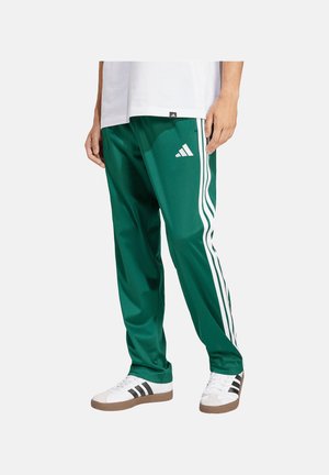 Hombre con pantalones de chándal verdes Adidas con tres rayas blancas y zapatillas blancas con rayas negras, de pie contra un fondo liso.
