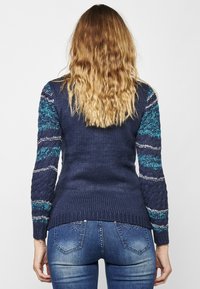 Marineblauer Pullover mit strukturierter Rückseite, farbenfrohen, gemusterten Ärmeln in Türkis und Weiß, geripptem Saum, getragen zu Skinny Jeans mit Verzierungen.