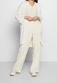 Žena v off-white blůze s širokým límečkem, volném cardiganě, kalhotách s vysokým pasem a širokými nohavicemi a bílých teniskách, stojící proti jednoduchému pozadí.