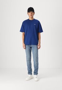 Blue cotton t-shirt with short sleeves, round neckline, and subtle logo. Paired with light blue jeans and white sneakers. Black cap worn.