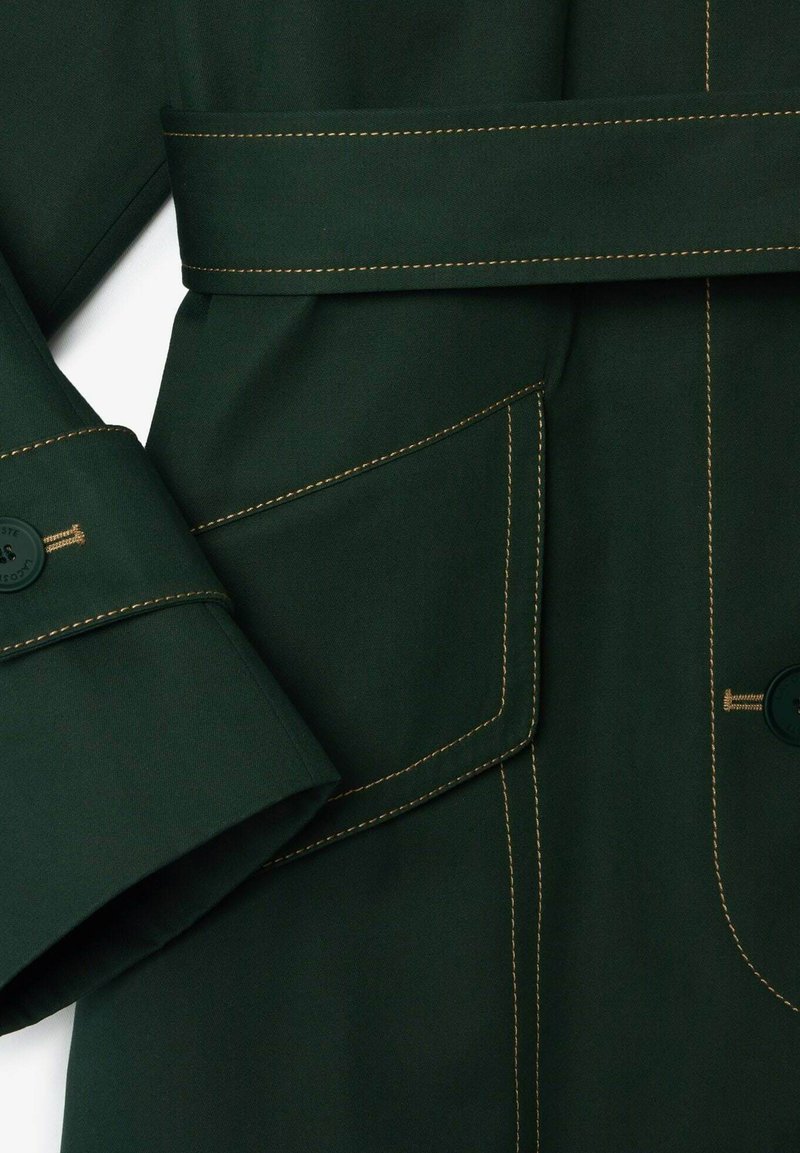 Manteau trench vert avec un col rabattu, des manches longues avec détails de boutons, des coutures dorées contrastantes, et une poche à l'avant.