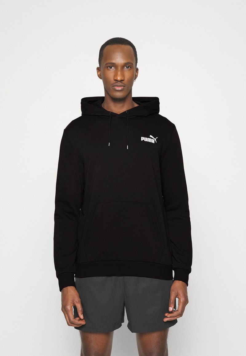 Czarna bluza z kapturem z kieszenią z przodu, logo PUMA w bieli. Miękka tkanina, długie rękawy, ściągacze przy mankietach i u dołu. Noszona z szarymi szortami.