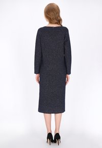 Sötétkék kötött ruha hosszú ujjakkal és kerek nyakkal, textúrált mintával és egyenes szabású, térd középig érő hosszúsággal. Fekete magassarkú cipő.