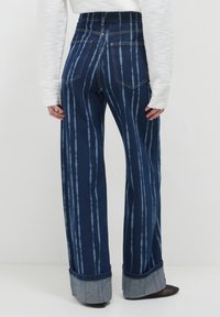 Persona che indossa jeans blu a vita alta e gamba larga, con strisce verticali di un azzurro chiaro e ampie risvolte, abbinati a un top bianco a maniche lunghe con texture.