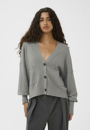 Šedý pletený cardigan s výstřihem do V, kleslými rameny, dvěma předními kapsami a tmavými knoflíky. Tkanina se zdá být měkká s hladkým povrchem.