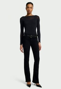 Woman standing wearing a fitted black long-sleeve top, black pants with a belt, and black pointed-toe heels against a plain background.