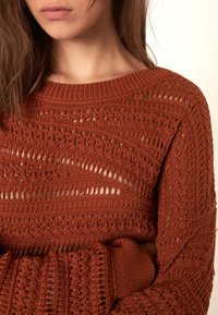 Rostfärgad stickad tröja med ett hålmönster, vid halsringning och långa ärmar med texturerade manschetter.