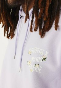 Sudadera con capucha de color púrpura claro, con bordados florales y texto blanco "PROJECT X PARIS GALERIE." Incluye cordones con puntas de metal.