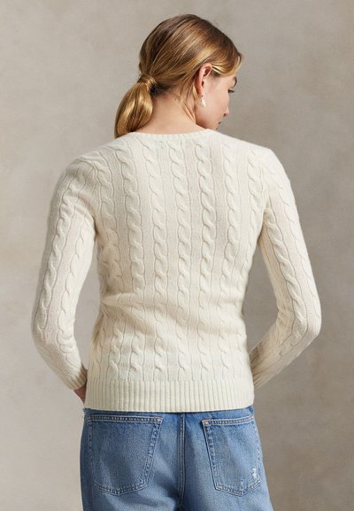 Femme aux cheveux châtain clair attachés en queue de cheval basse, portant un pull en maille torsadée crème et un jean bleu, vue de dos sur un fond neutre.