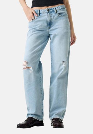 Jeans strappati azzurro chiaro indossati con mocassini neri e top nero corto, mano nella tasca, su sfondo semplice.