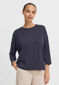 Dunkelblaues Langarmshirt mit rundem Ausschnitt und lockerer Passform. Weicher Stoff, entspannte Ärmel und sichtbare Nähte. Kombiniert mit hellen Hosen.