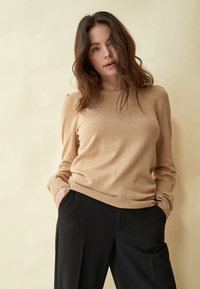 Beige neulepaita, jossa on pitkät hihat, ribbaatut hihansuut ja helma, yhdistetty mustiin housuihin. Pehmeä rakenne, rento istuvuus ja hienoinen kiilto.